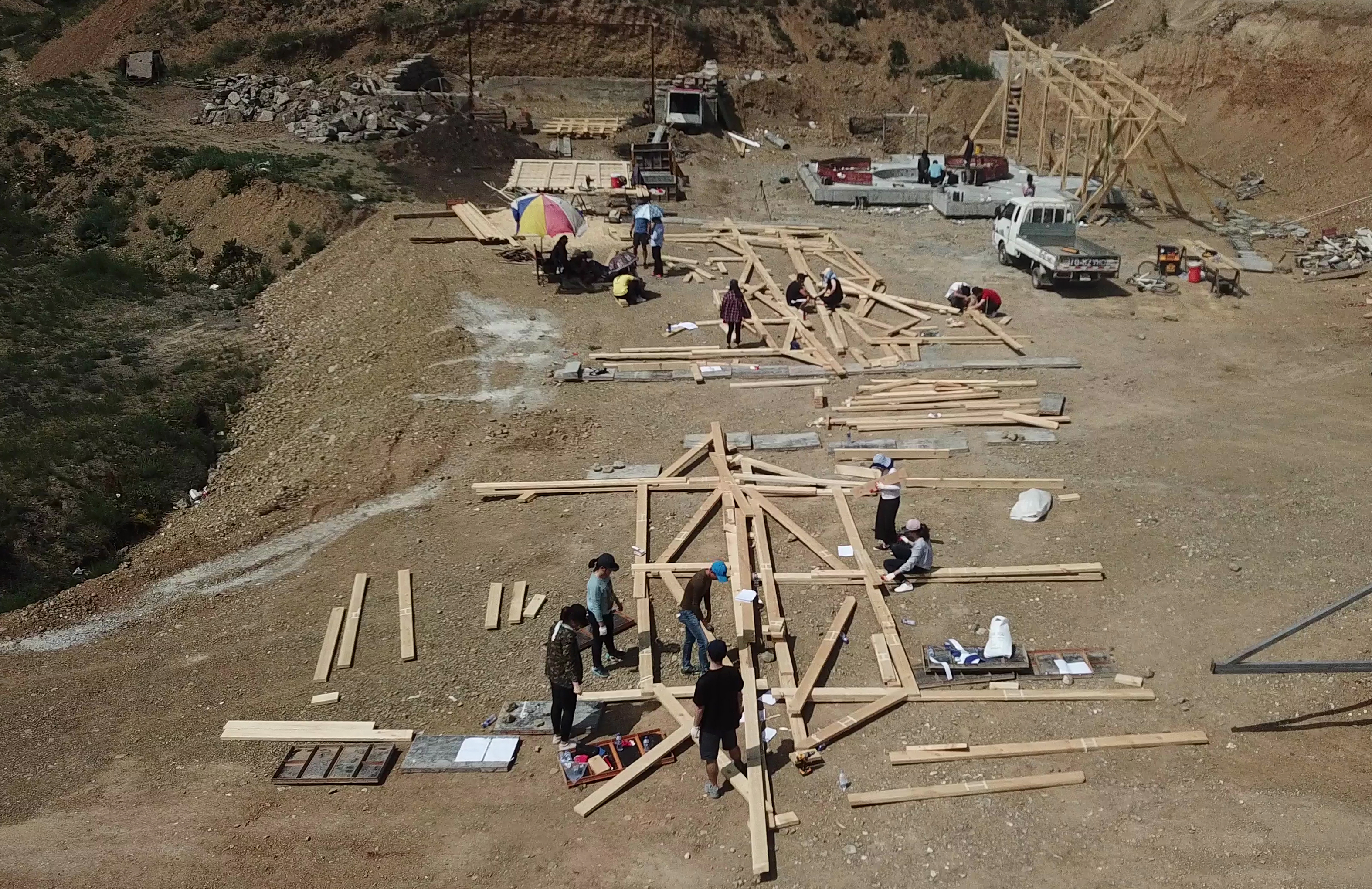 Construction site with wooden beams laid out on a dirt plain. People gathered around discussing the project. 